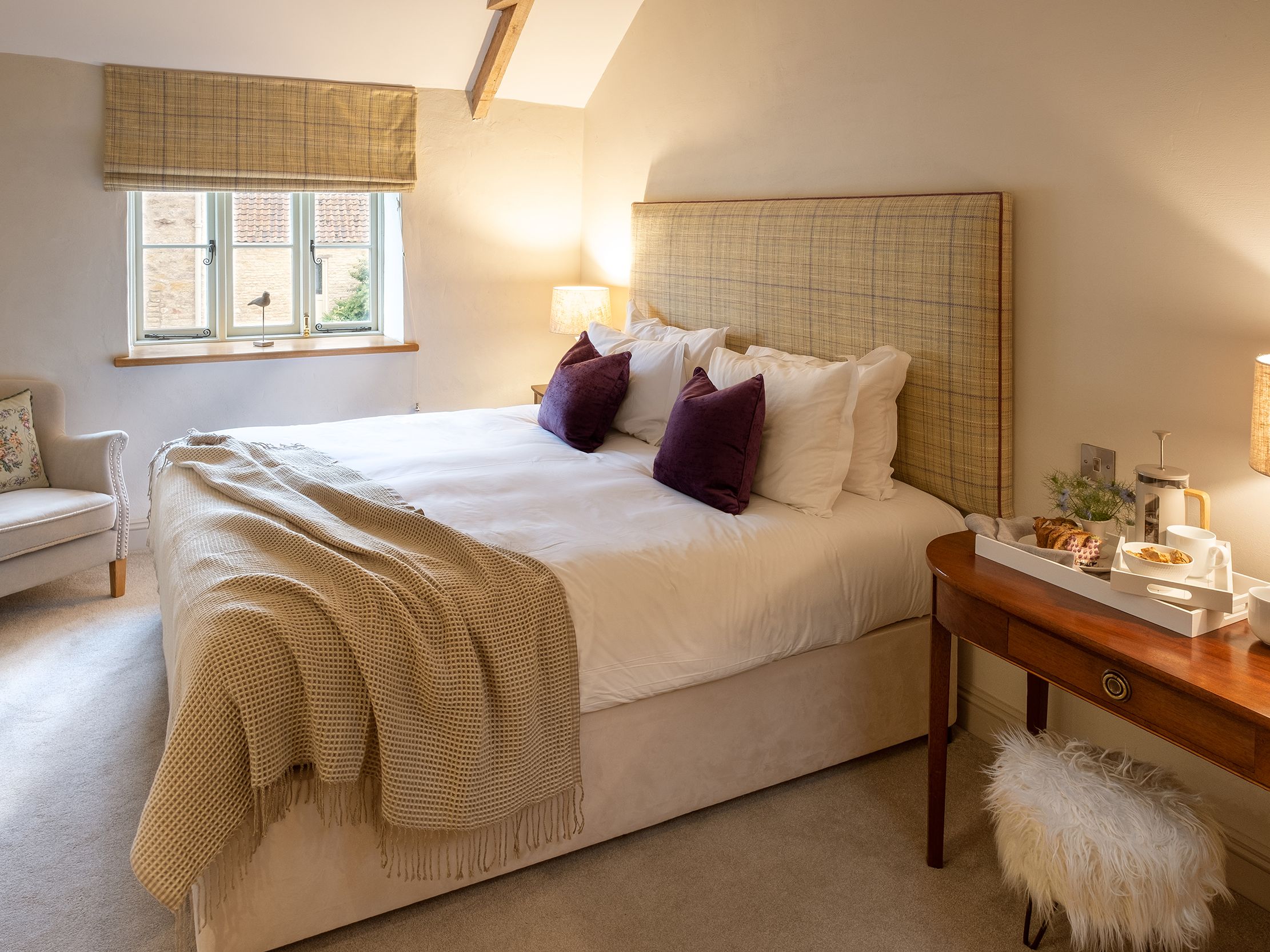 Cozy bedroom with a large bed, two purple cushions, a beige blanket, and a wooden side table with breakfast tray and lamp.