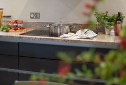 Modern kitchen countertop with a pot on a stove, fresh herbs, tomatoes, pasta jars, olive oil, and cooking utensils.