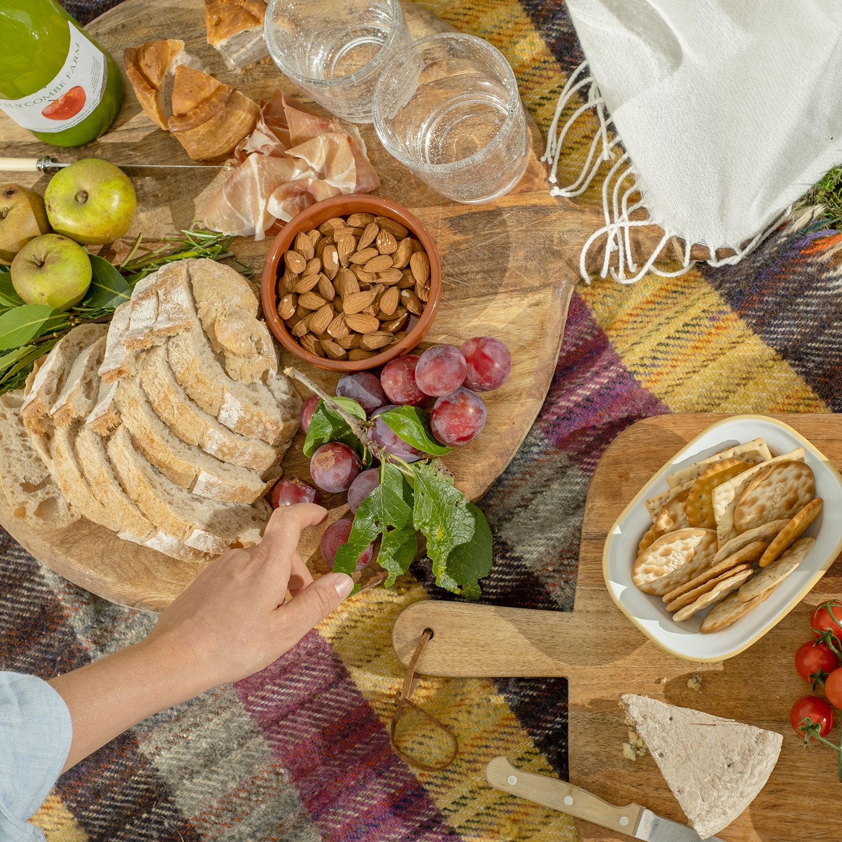 A picnic spread on a plaid blanket with bread, apples, grapes, almonds, crackers, cherry tomatoes, preserves, and a bottle of juice.