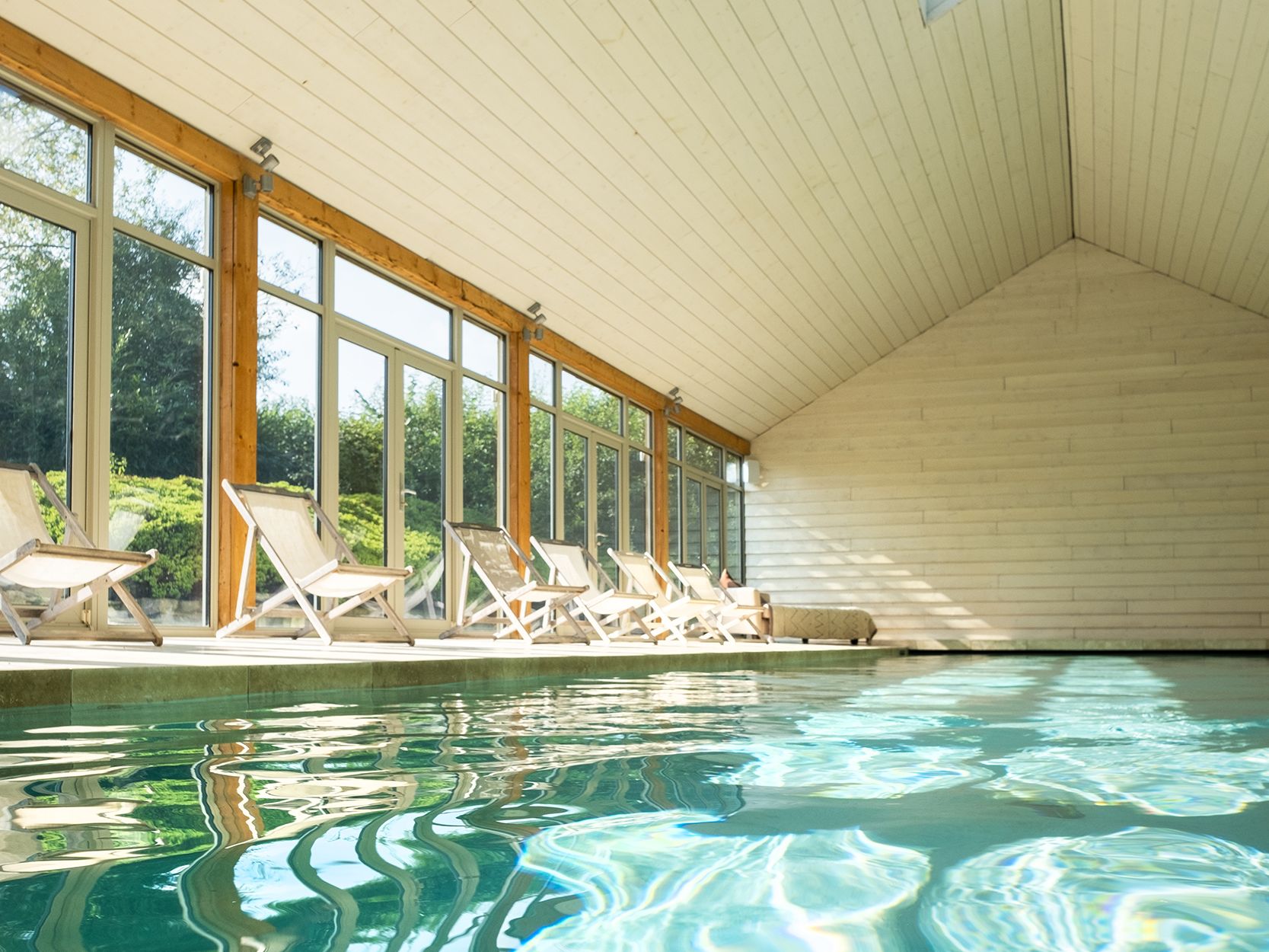 Indoor swimming pool with wooden ceiling, skylight, and lounge chairs by large windows.
