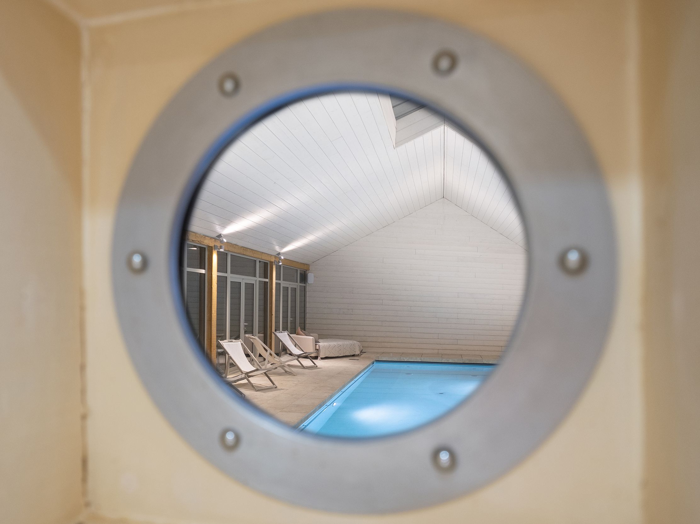 View through a porthole window into an indoor swimming pool with lounge chairs.