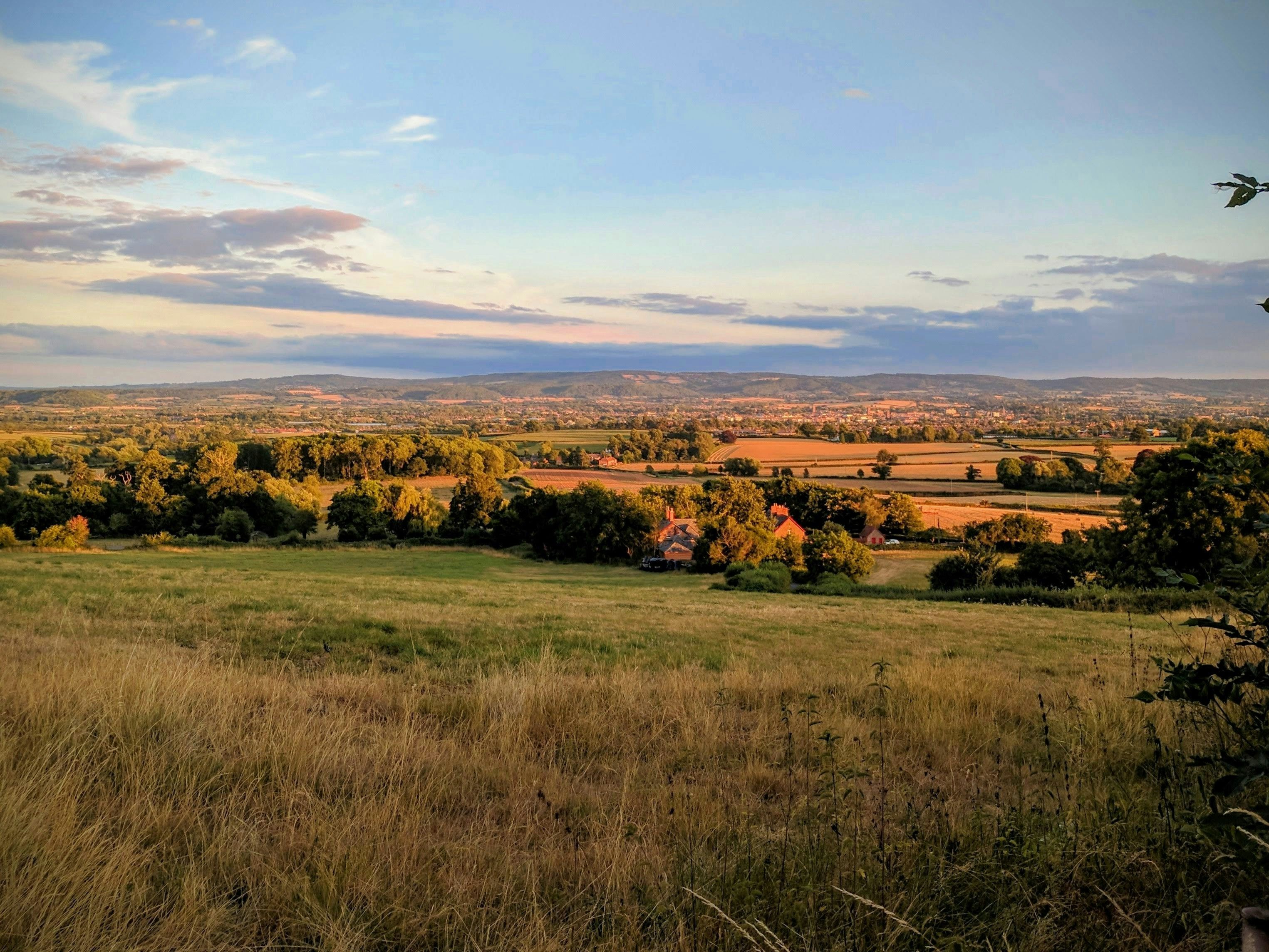 A scenic rural landscape with rolling fields, green trees, and distant houses under a partly cloudy sky at sunset.