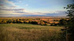 A scenic rural landscape with rolling fields, green trees, and distant houses under a partly cloudy sky at sunset.