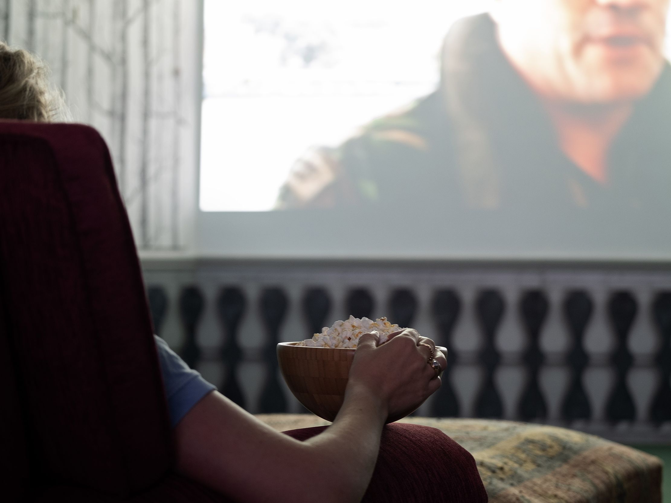 Person sitting on a chair holding a bowl of popcorn while watching a movie on a large screen.