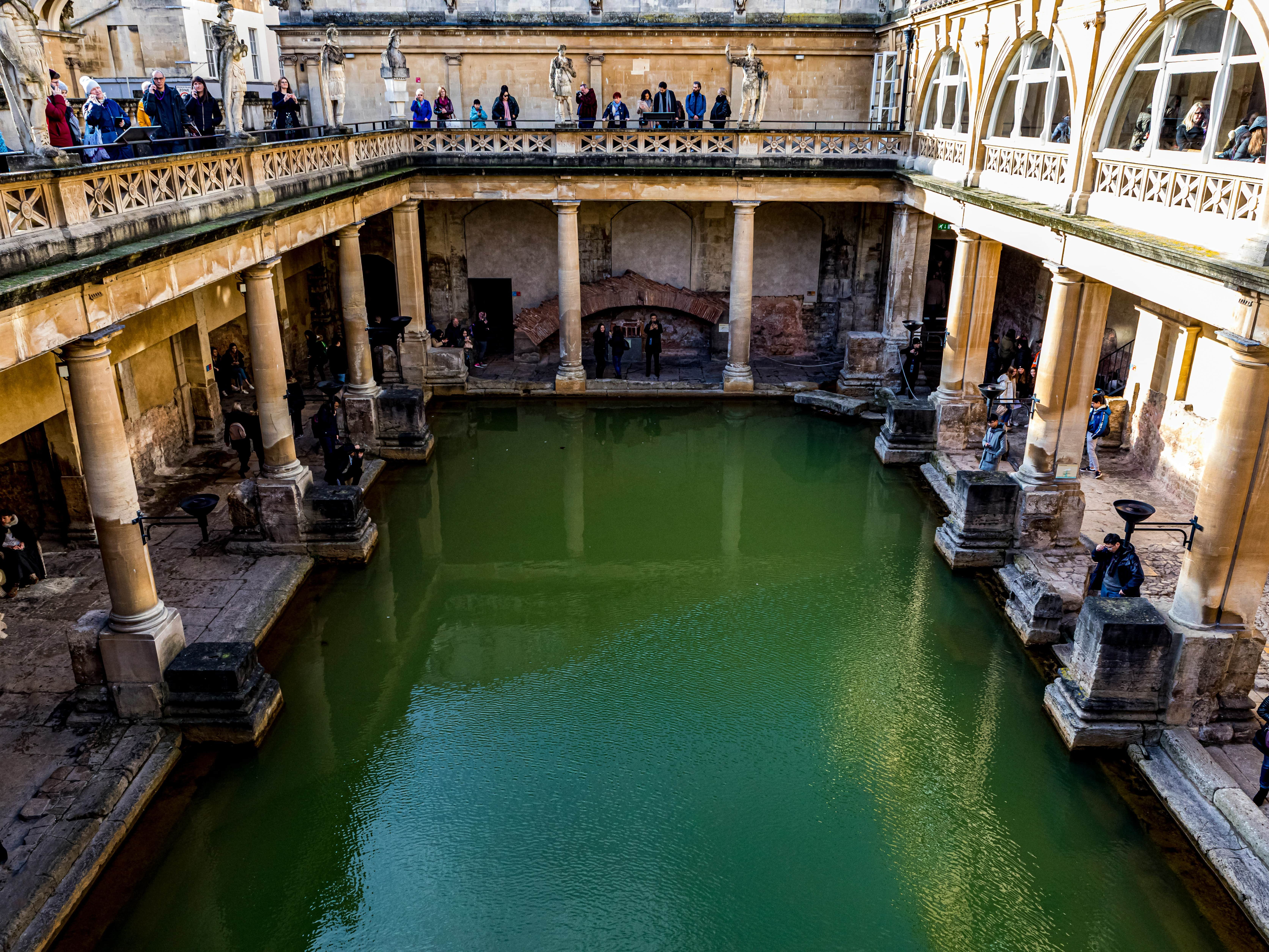 Ancient Roman baths with green water surrounded by columns and people walking along the paths.