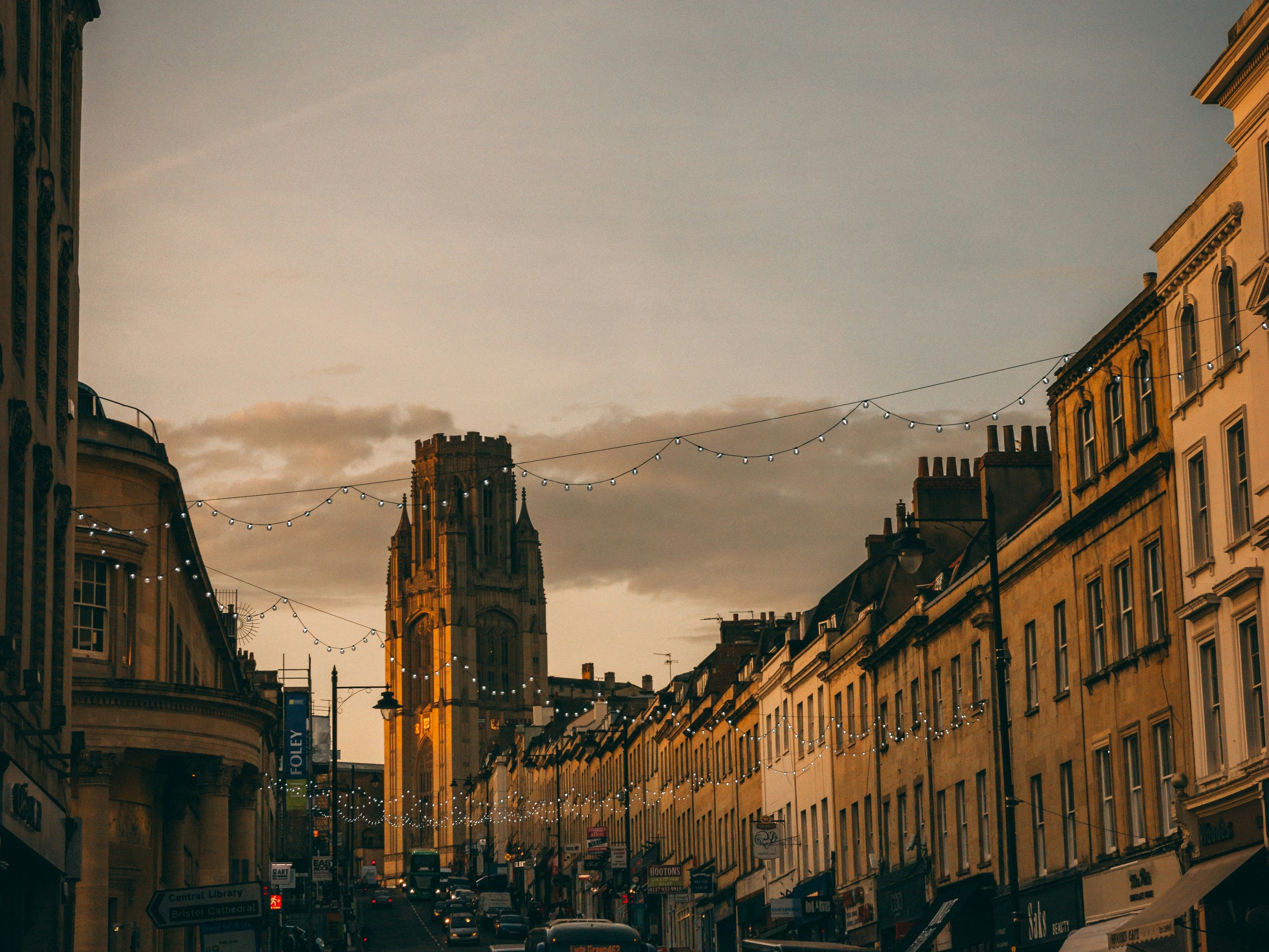 A city street at sunset with cars, shops, and a tall historic tower in the background.