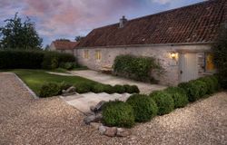 Charming stone cottage with a red tile roof, lit exterior at dusk, with a landscaped garden path and bench.