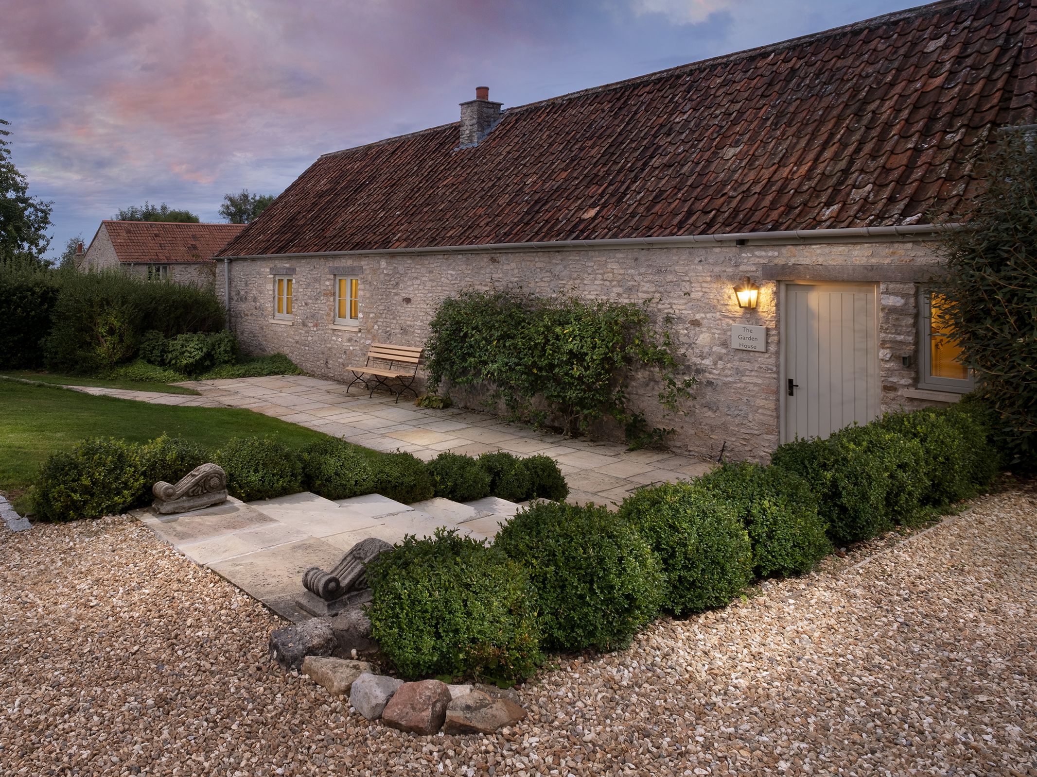 Charming stone cottage with a red tile roof, lit exterior at dusk, with a landscaped garden path and bench.