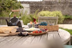 Outdoor breakfast table set with bread, fruit, a kettle, and plates.