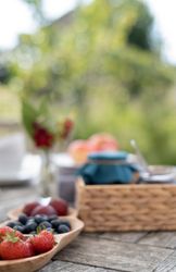 Close-up of a wooden tray with strawberries, blueberries, and plums on an outdoor table with jars and blurred greenery in the background.