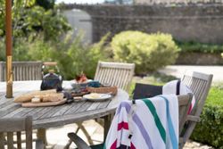 Outdoor wooden table set for a meal with bread, cheese, and tea, surrounded by chairs with striped towels draped over them.