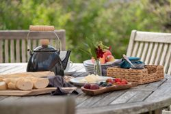 Outdoor table set for breakfast with a teapot, bread, fruit, and jam.