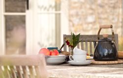 Outdoor table set with a black kettle, apples on plates, cups, and a small flower vase