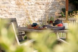 Outdoor wooden table with chairs, fruit, a teapot, flowers, and cups set for breakfast or tea.