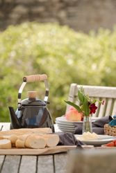 Outdoor breakfast or picnic setup with black kettle, bread, butter, fruit, plates, and flowers on a wooden table.