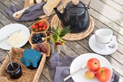 Outdoor breakfast setup with tea kettle, fruits, bread, butter, jam, apples, and cups on a wooden table