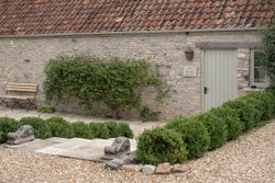 Stone cottage exterior with a sign reading 'The Garden House', green bushes, a bench, and a gravel path.