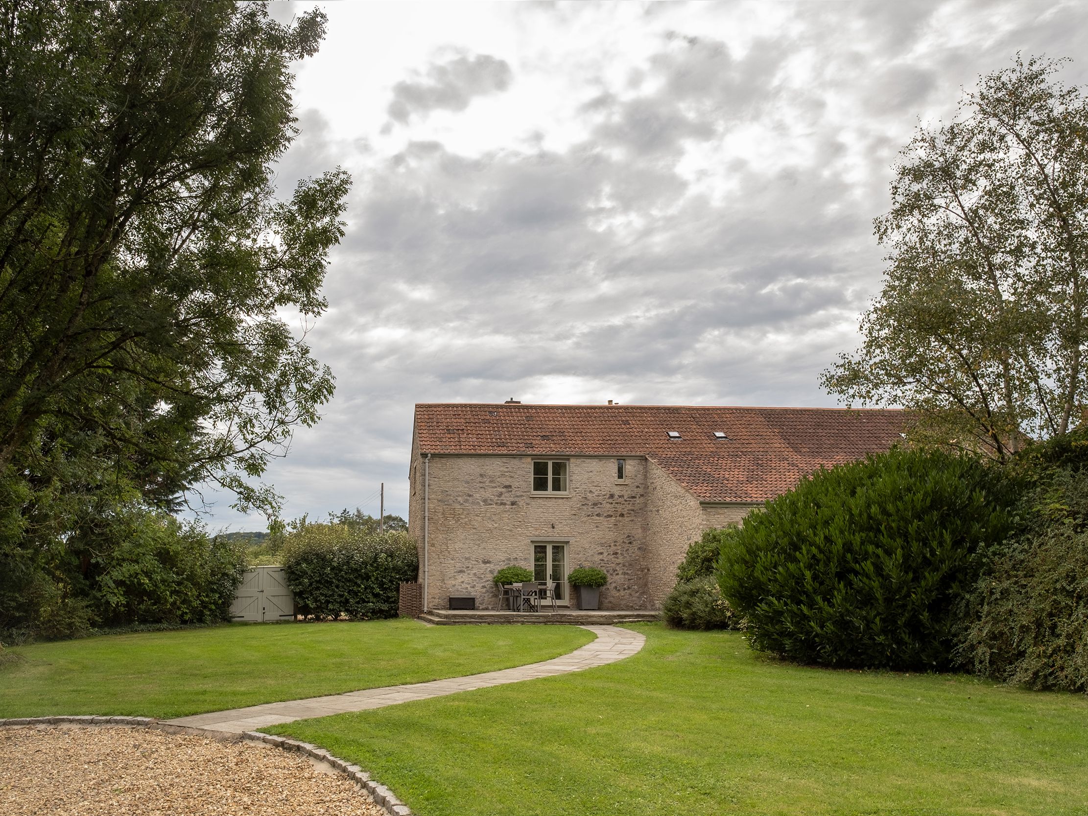 Stone house with red tiled roof surrounded by green lawn and trees under cloudy sky