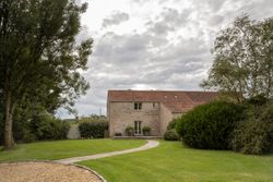 Stone house with red tiled roof surrounded by green lawn and trees under cloudy sky