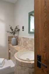 Small bathroom with a tiled sink and countertop, a gray vase with green leafy branches, a mirror with a green frame, and a wooden door partially open.