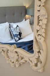 Reflection of a cozy bed with white pillows, a decorative cushion, and a grey headboard seen through an ornate mirror