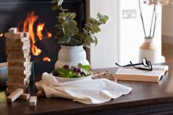 Table with a Jenga game, bowl of grapes, a book with glasses, and a vase of greenery in front of a fireplace.