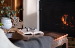 A cozy scene with an open book and eyeglasses on a wooden table in front of a fireplace.