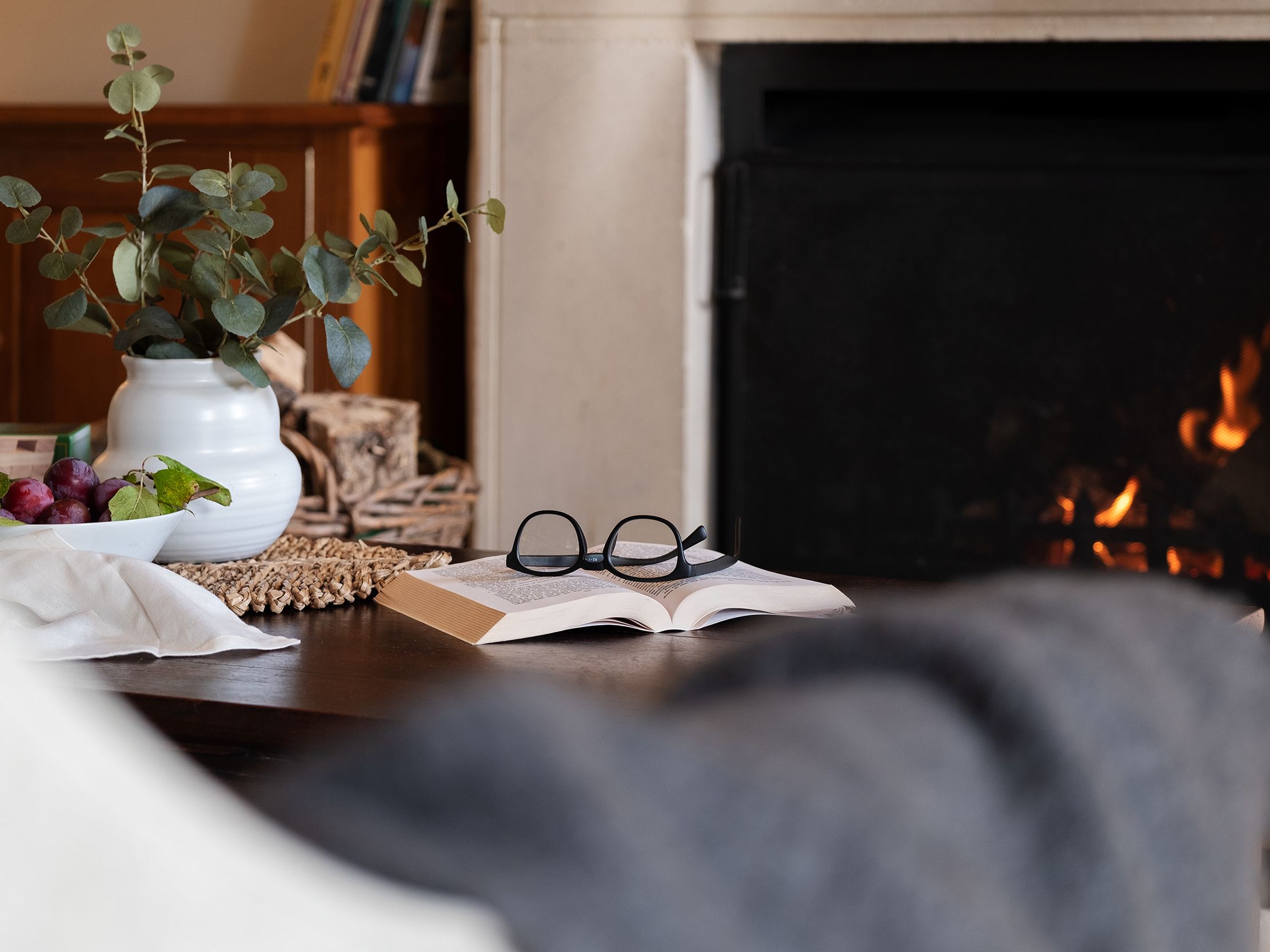 Cozy living room scene with a book, glasses, a vase with greenery, a fruit bowl, and a fireplace.
