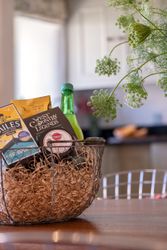 Gift basket with snacks, tea, a drink, and cheese on a wooden table with flowers in the background