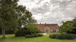 A country house with a red-tiled roof surrounded by a lush green garden and large trees under a cloudy sky.