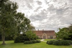 A country house with a red-tiled roof surrounded by a lush green garden and large trees under a cloudy sky.