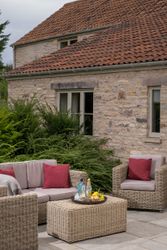 Outdoor patio with wicker furniture, red cushions, and refreshments on a table in front of a stone house.