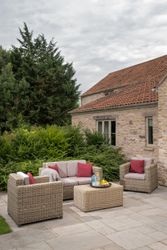 Outdoor wicker patio furniture with red cushions and a coffee table in a garden setting next to a stone house.