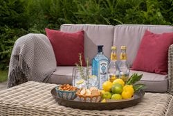 Outdoor table with gin, tonic water, snacks, lemons, and herbs on a tray in front of a wicker sofa with red pillows.