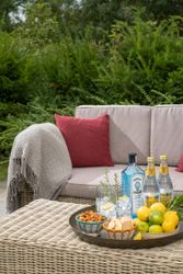 Outdoor wicker table with a tray of Bombay Sapphire gin, tonic water, snacks, lemons, and limes near a cozy sofa with red pillows and throw blanket.