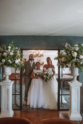Bride, groom, and a young bridesmaid posing together indoors between two large floral arrangements.