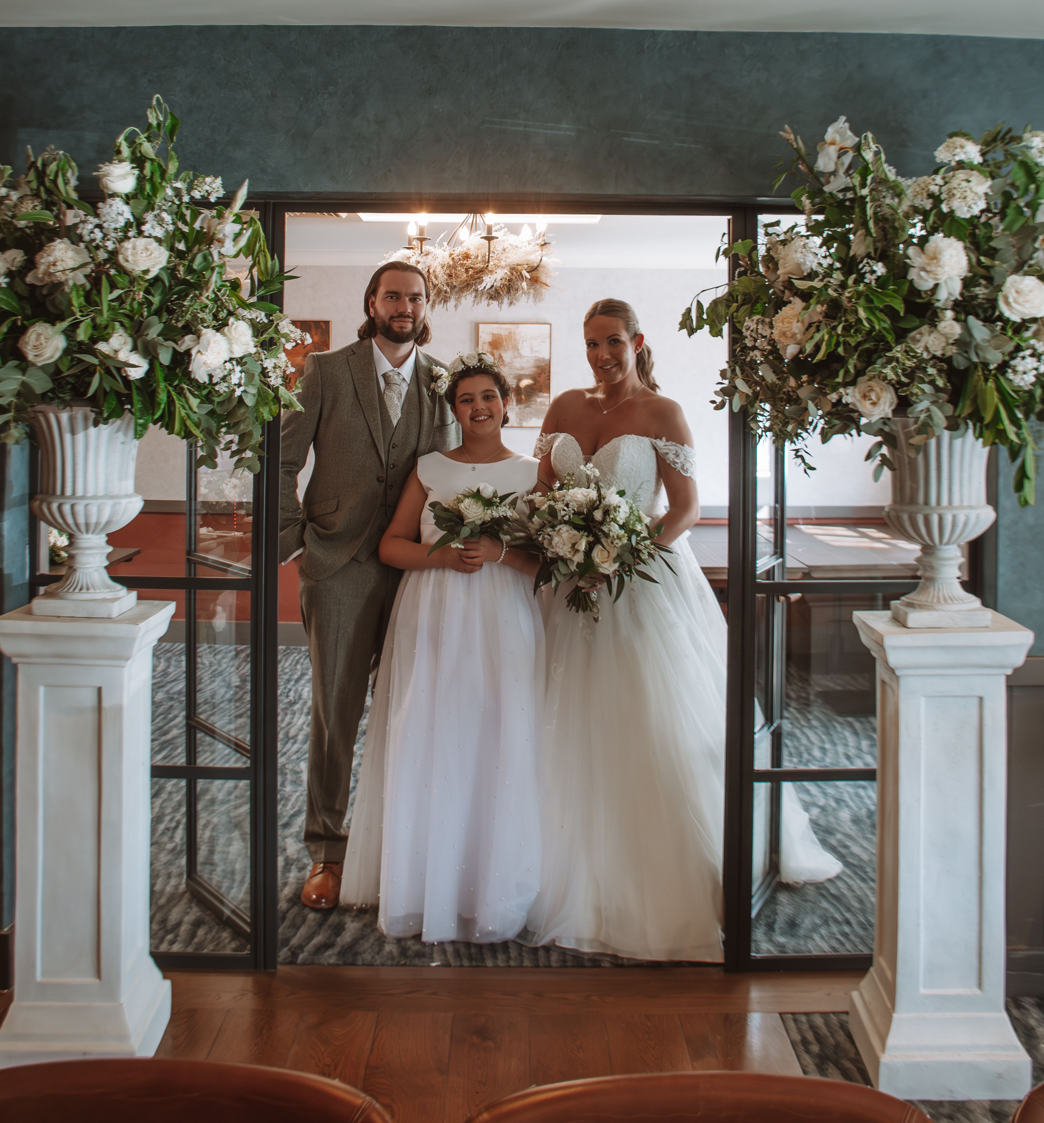 Bride, groom, and a young bridesmaid posing together indoors between two large floral arrangements.