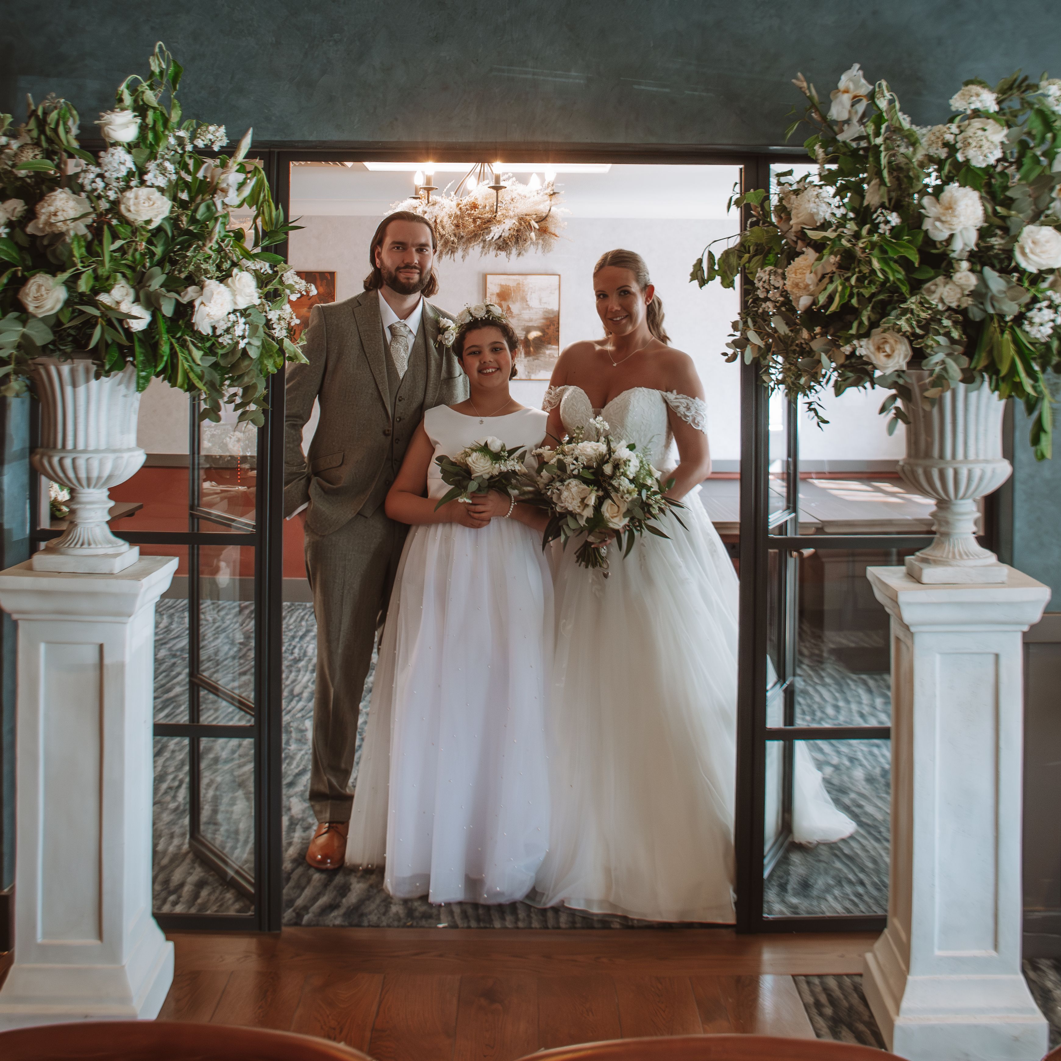 Bride, groom, and a young bridesmaid posing together indoors between two large floral arrangements.