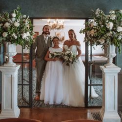 Bride, groom, and a young bridesmaid posing together indoors between two large floral arrangements.