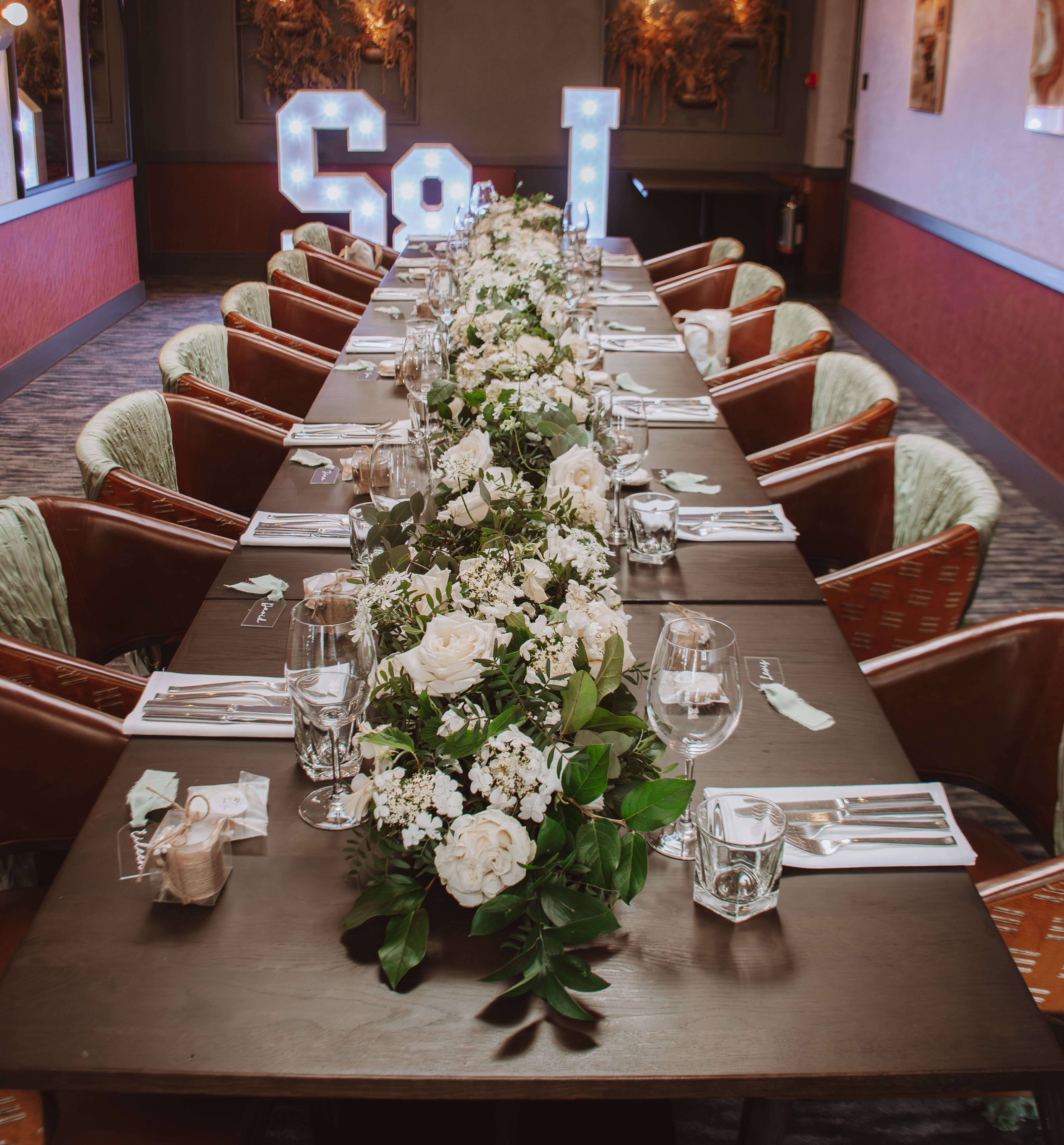 Elegant dining table set for an event, decorated with white flowers and greenery, with illuminated letters in the background.