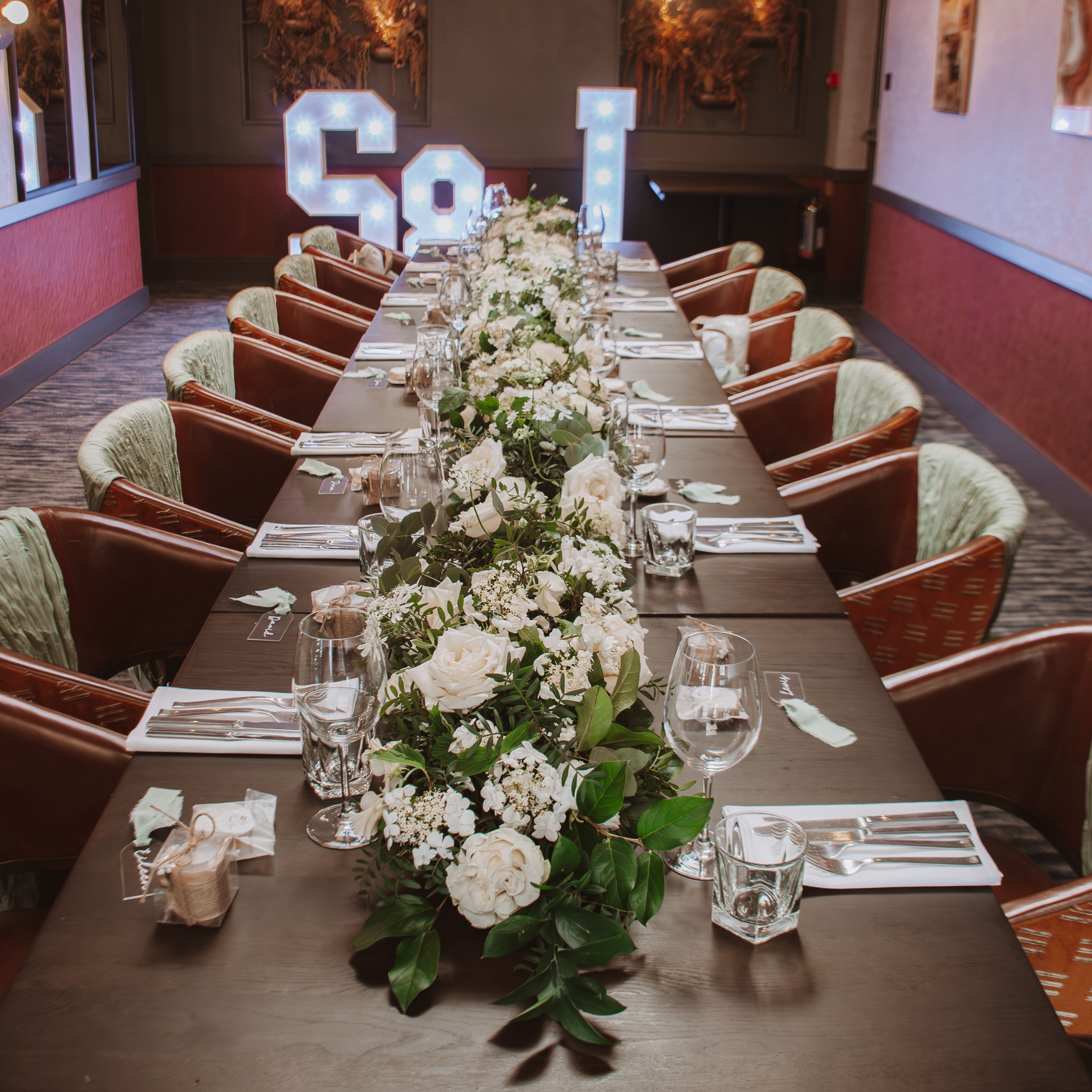 Elegant dining table set for an event, decorated with white flowers and greenery, with illuminated letters in the background.