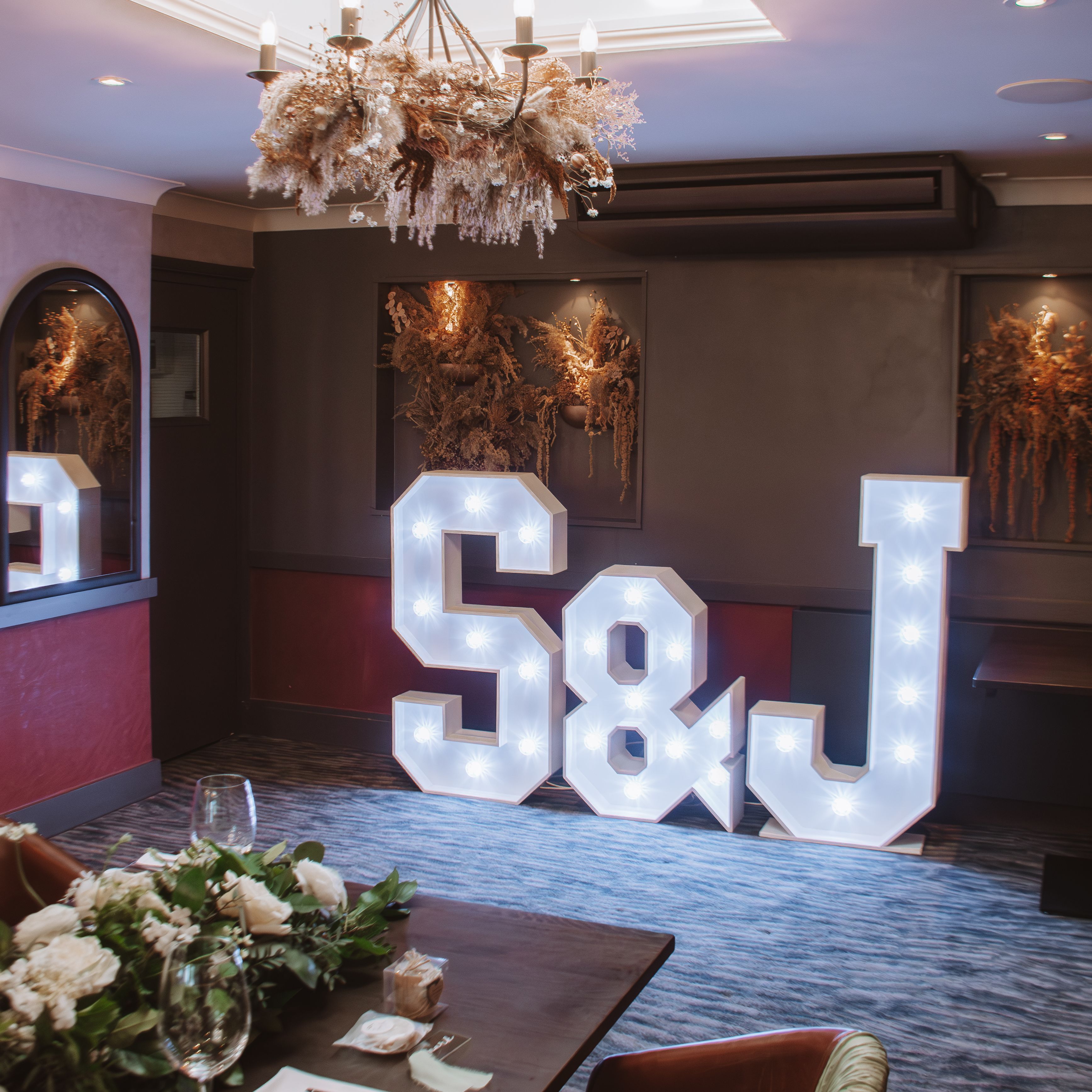 Illuminated large letters S & J in a decorated room with floral arrangements and a chandelier above.