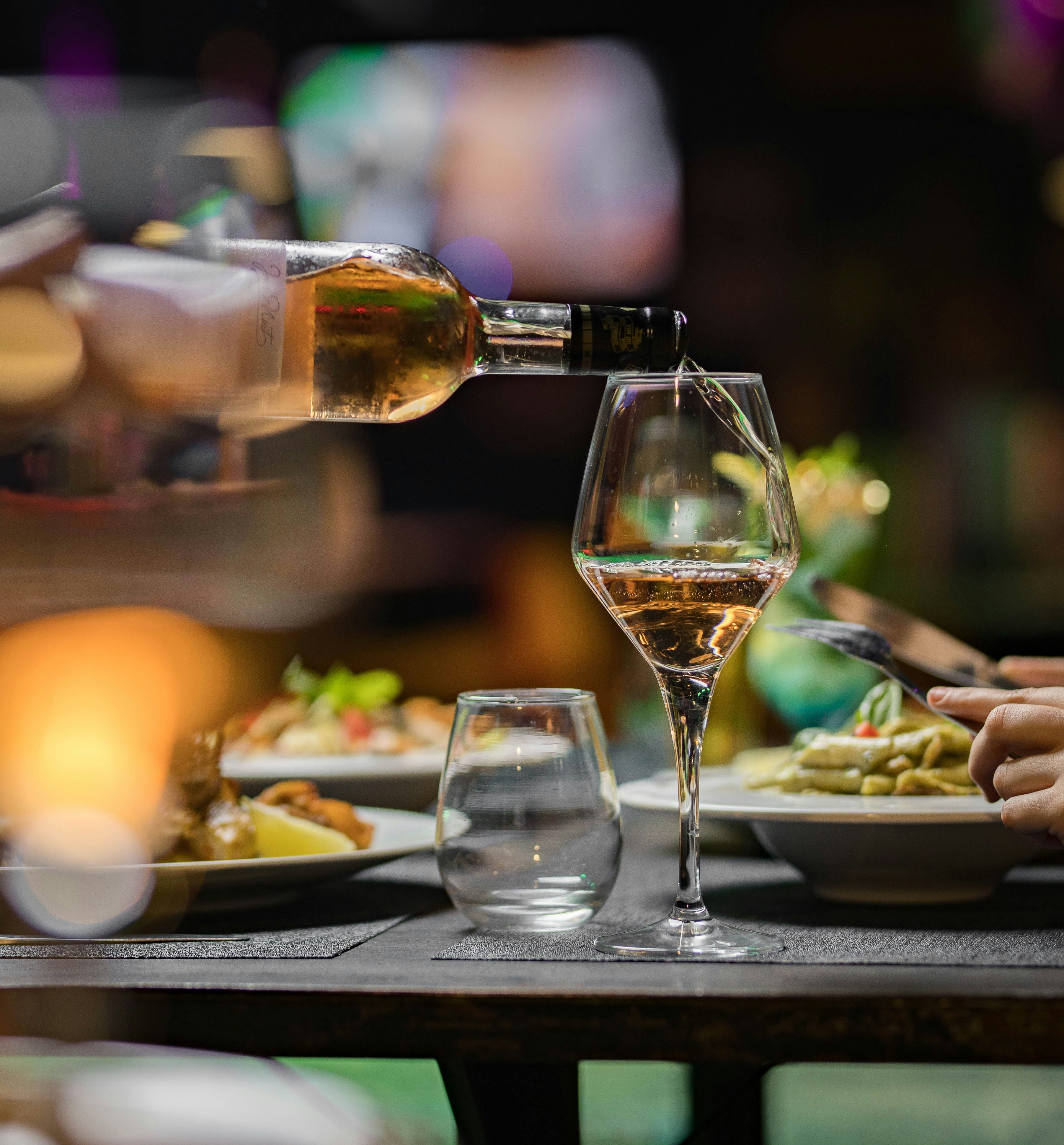 A bottle of white wine being poured into a wine glass at a restaurant table with plates of food.
