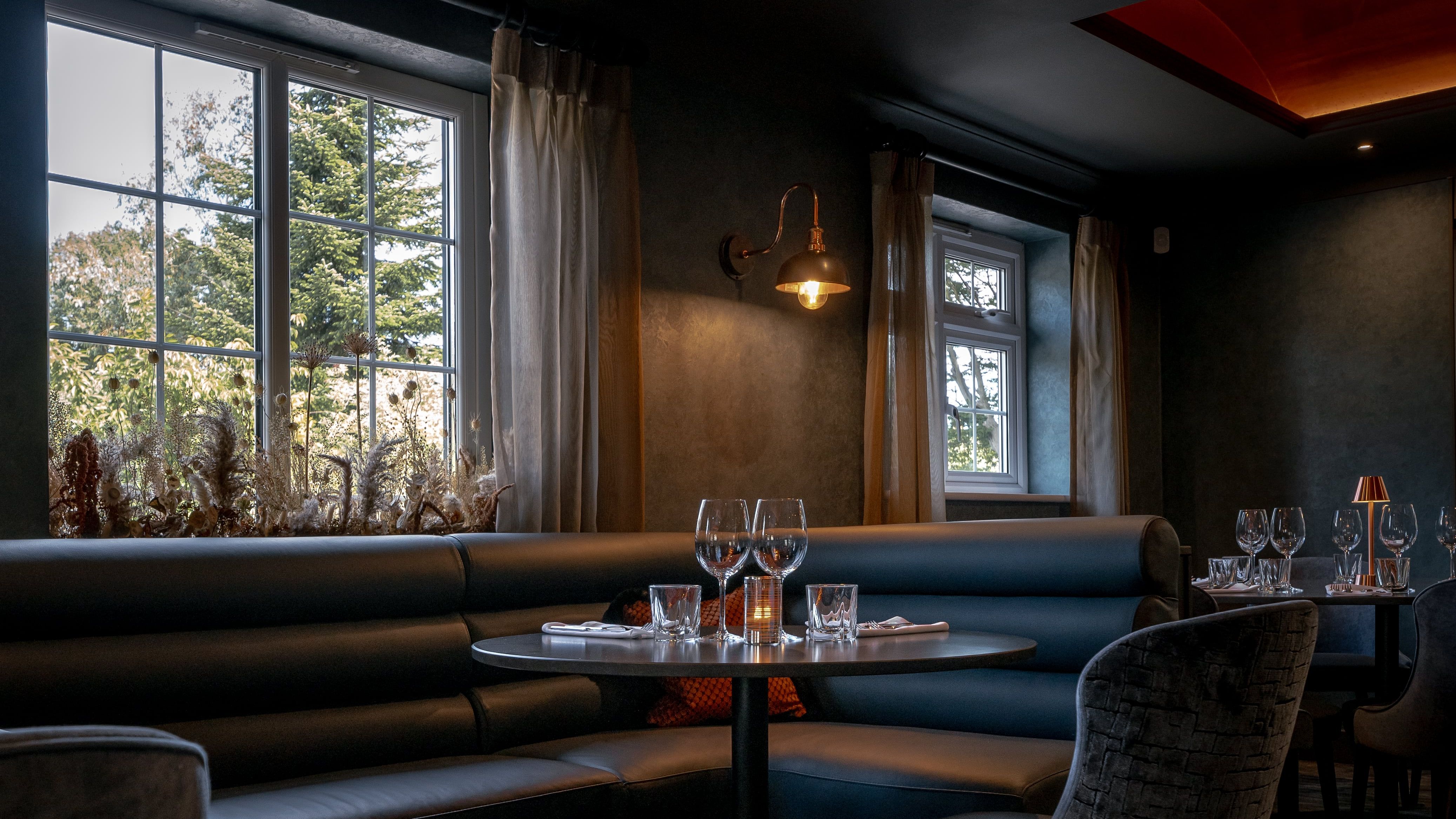 Elegant restaurant interior with a round table, cushioned booth seating, chairs, and set dining tableware near large windows.