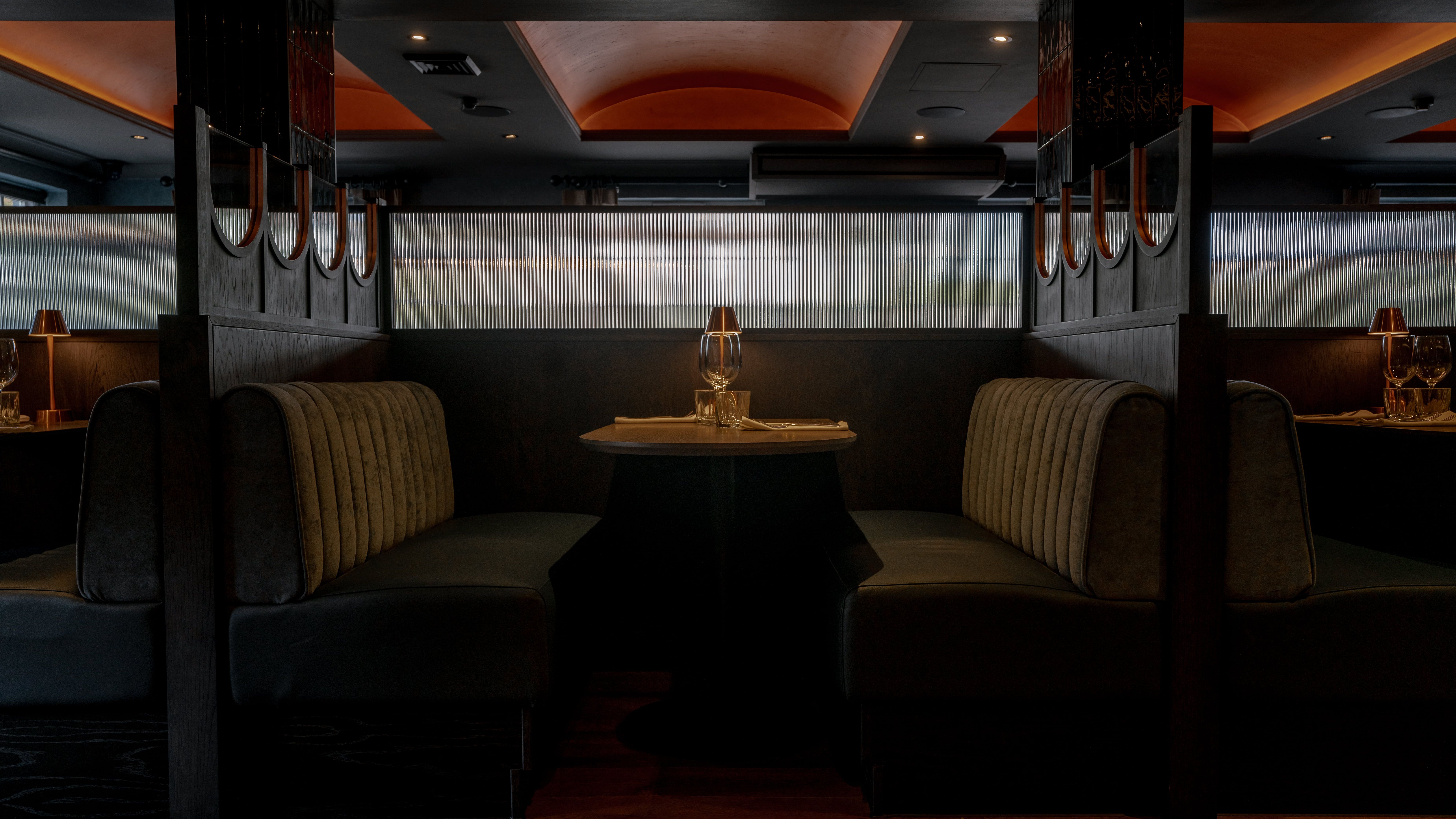 Modern restaurant booth seating with table set for two, softly lit by a small lamp
