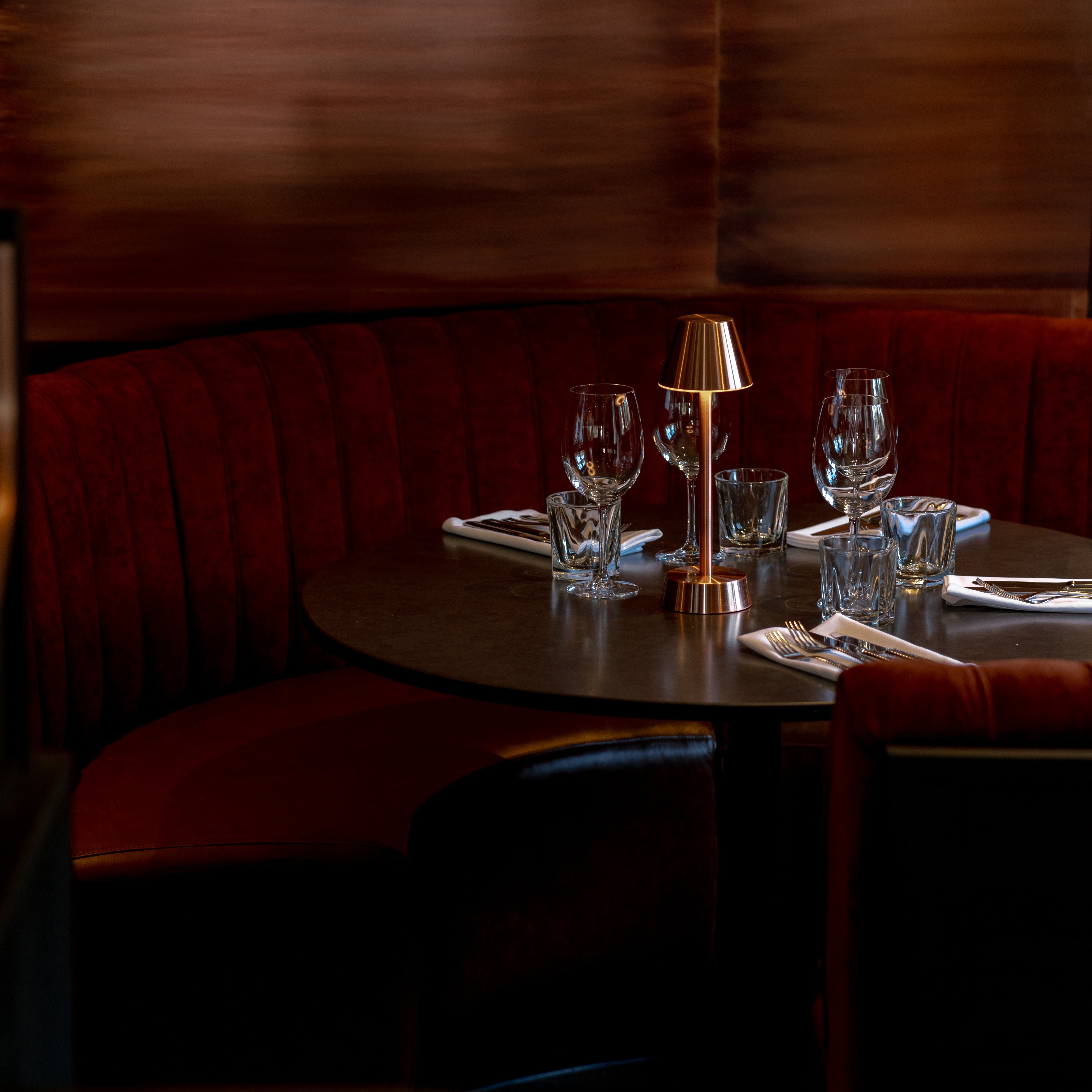Elegant restaurant booth with red velvet seating, set tableware, and a small golden lamp.