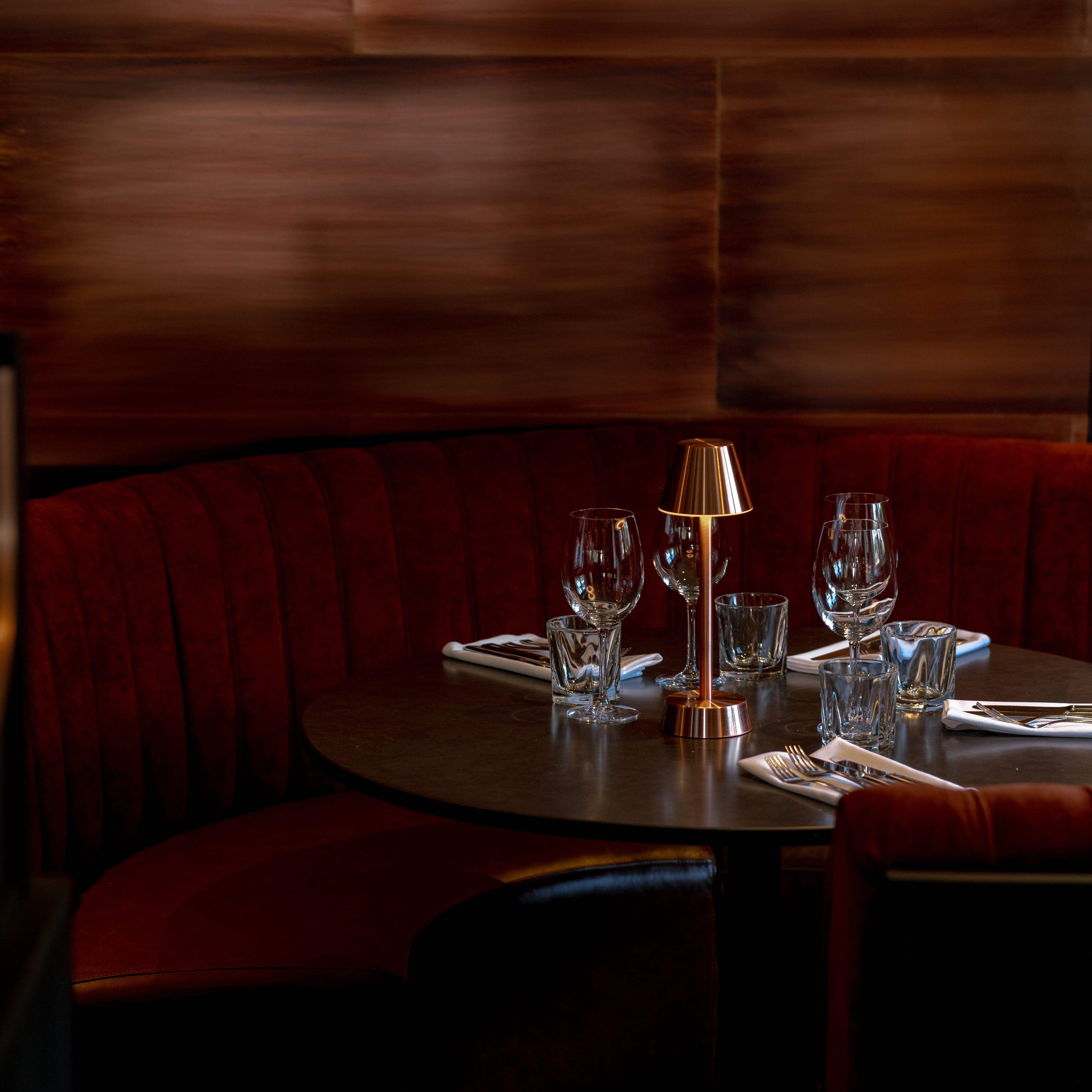 Elegant restaurant booth with red velvet seating, set tableware, and a small golden lamp.
