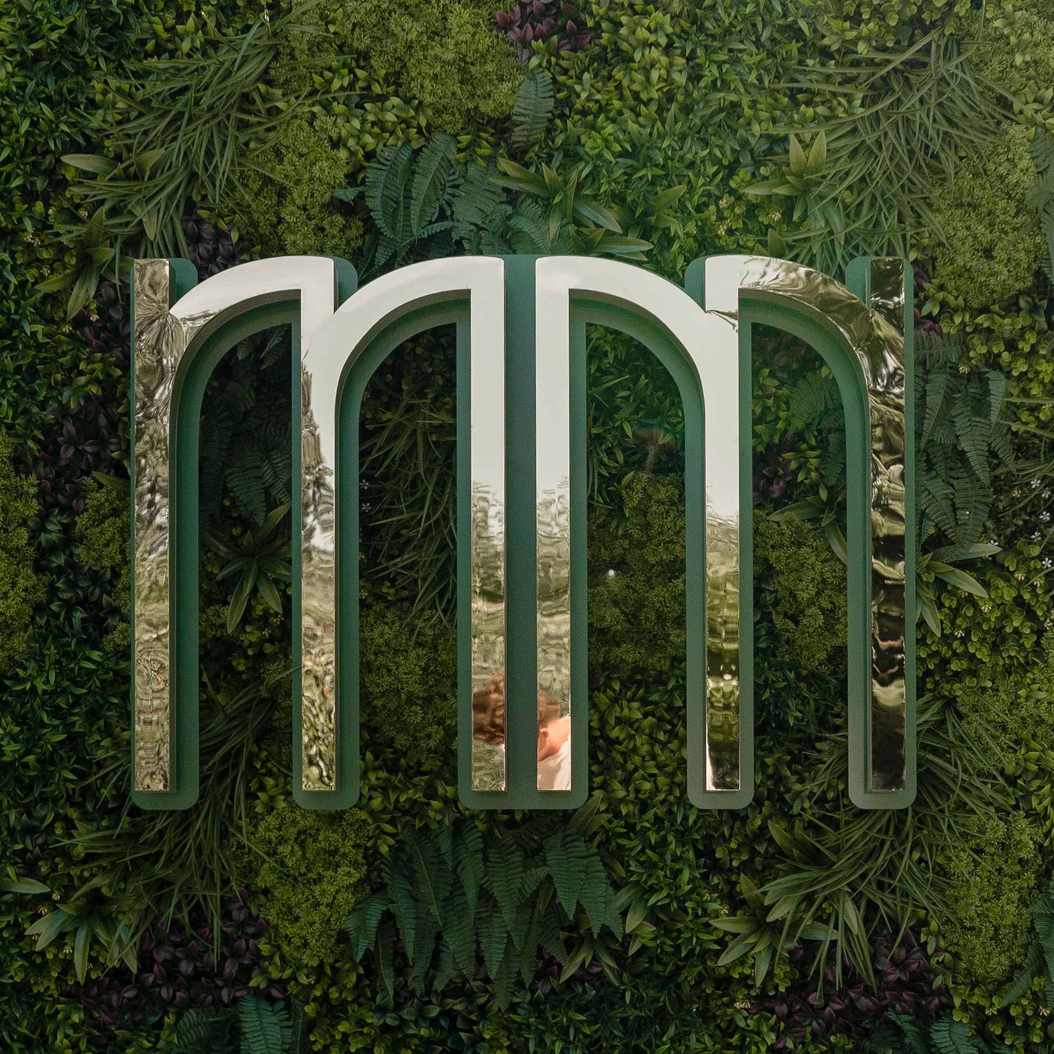 Large metallic 'mm' sculpture mounted on a lush green plant wall