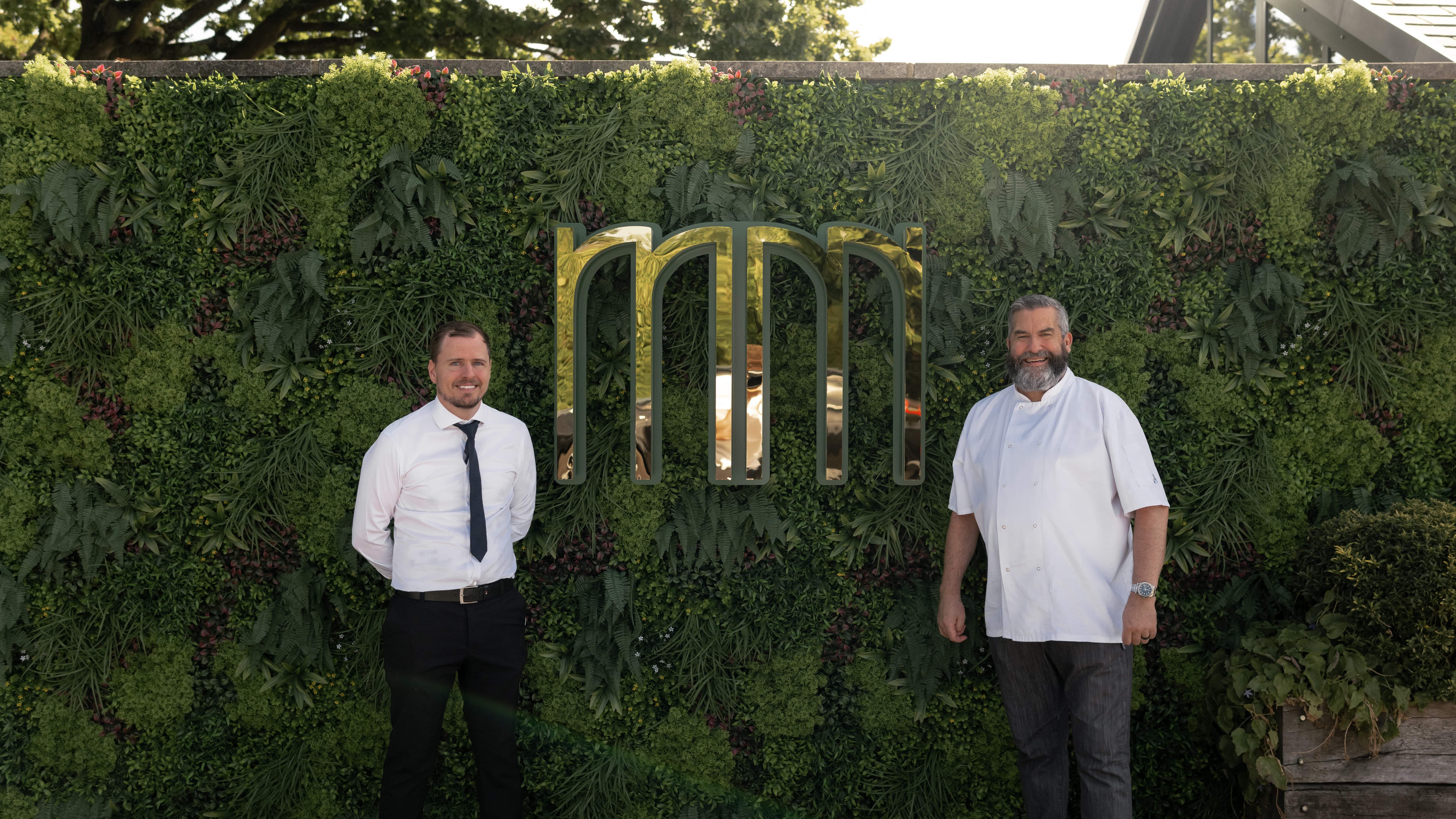 Two men standing in front of a green wall with a stylish logo, one in a shirt and tie, the other in a chef's coat.