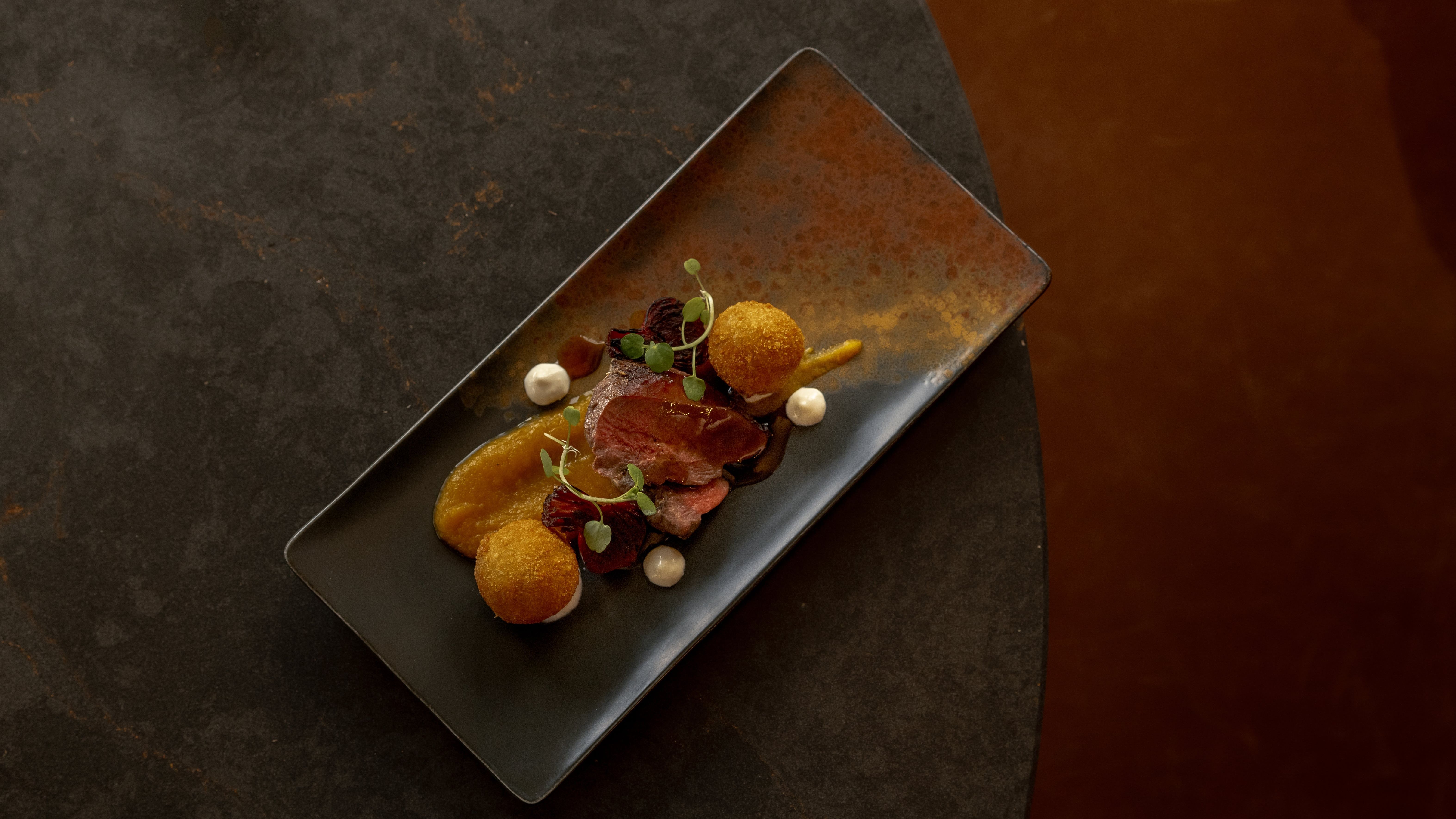 A gourmet plated dish with sliced meat, sauces, microgreens, and two golden-fried balls on a rectangular plate.